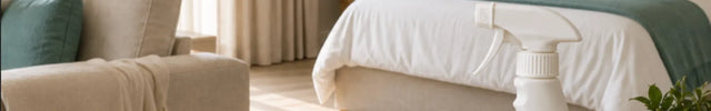 A bottle of Olorsol Professional Smoke Odor Eliminator placed on a round wooden table in a beautifully styled, sunlit bedroom. The room features a beige sofa, a bed with white and teal bedding, a large potted palm, and a natural jute rug.